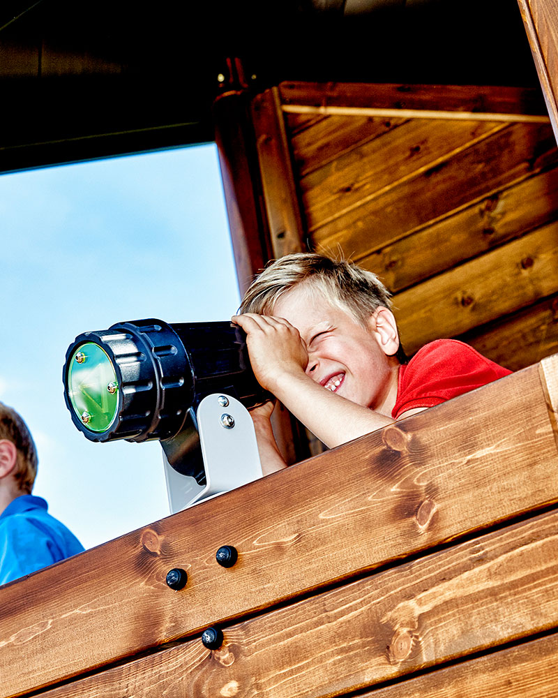 Young boy looks through a telescope on top of a playground system which is wooden.